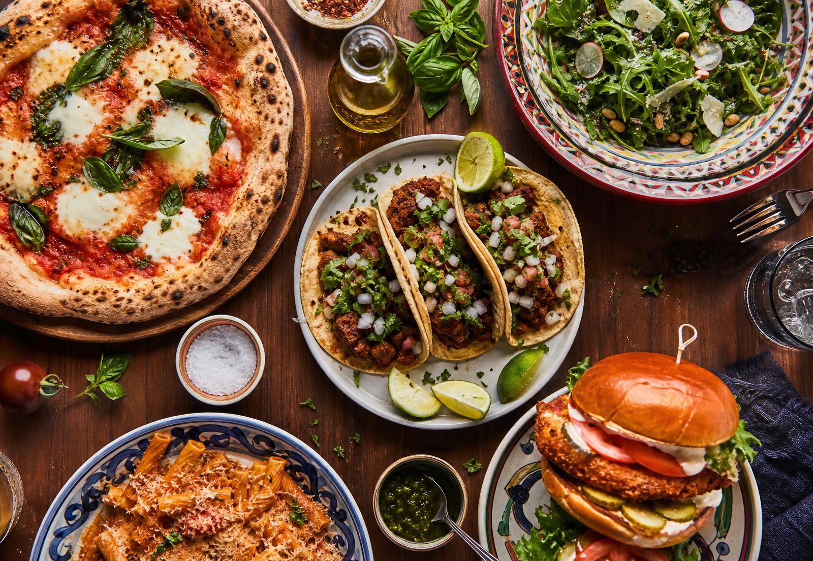 Overhead spread of homemade pizza, tacos, sandwiches and pasta dishes